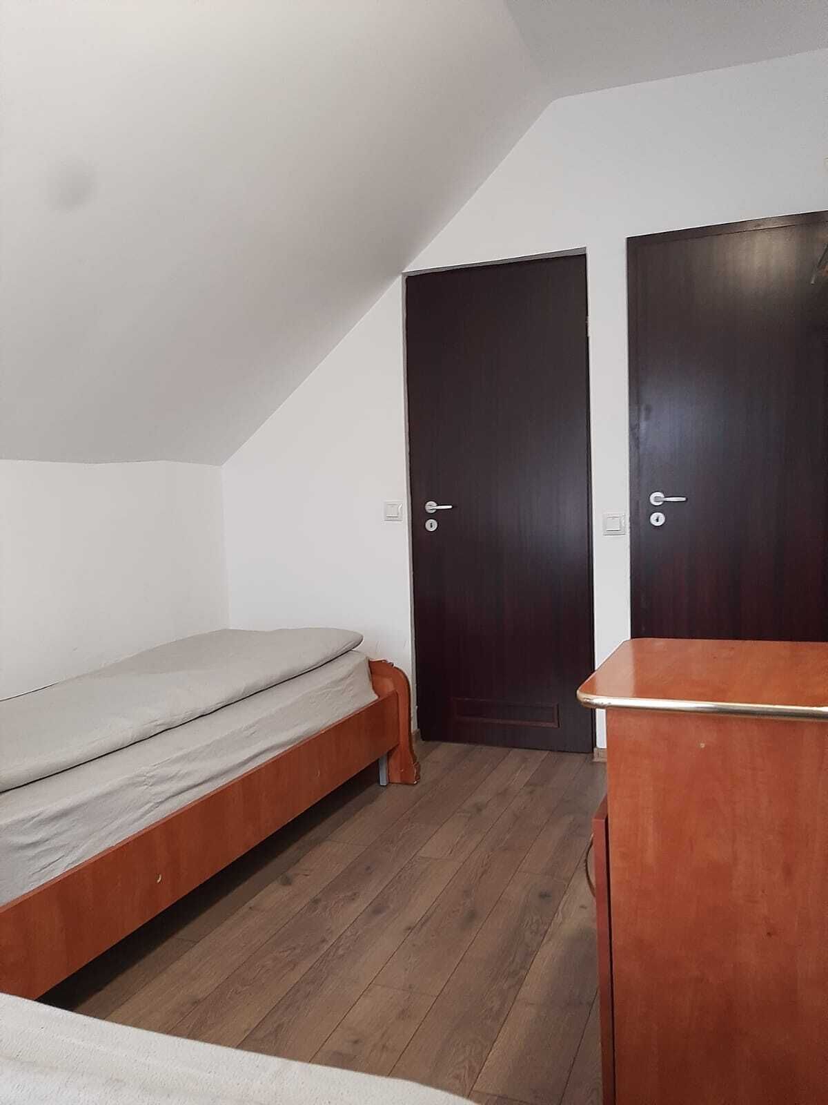 Eine kleine Einzimmerwohnung mit schrägem Dach, Holzboden, einem Bett, zwei dunklen Holztüren und einem kleinen Holztisch.
