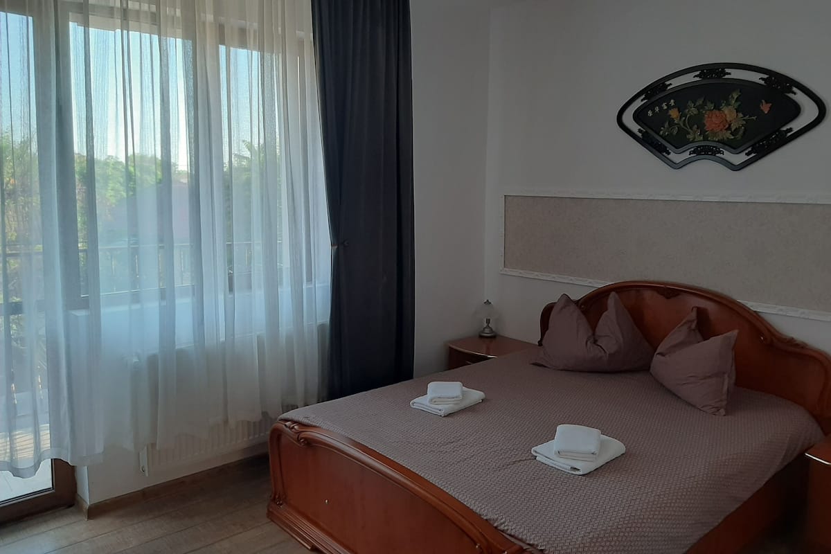 Ein Schlafzimmer mit einem Bett, Kissen, einem Nachttisch, einer Lampe, einem Fenster mit durchsichtigen Vorhängen und einer Wanddekoration.