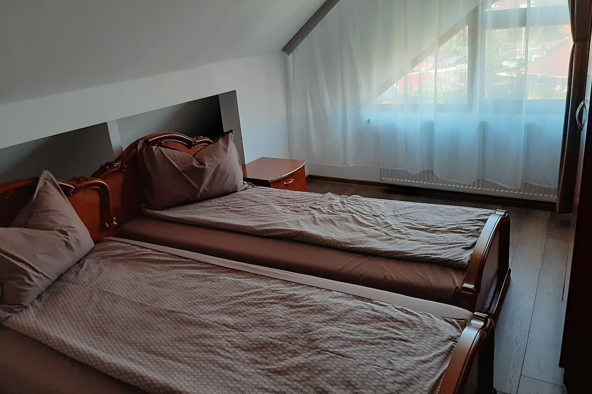 Ein Zimmer mit zwei Betten, einem Nachttisch und einem Fenster mit durchsichtigen Vorhängen.