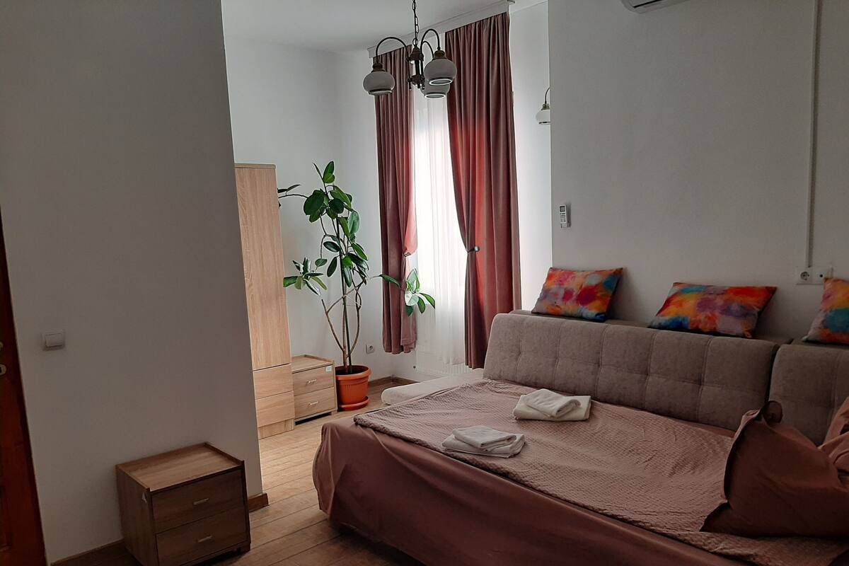 Ein Schlafzimmer mit Bett, Nachttisch, Kommode, einer Topfpflanze und einem Fenster mit Vorhängen.