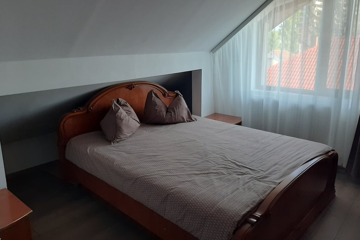 Ein Schlafzimmer mit einem hölzernen Bett, zwei Nachttischen, einem Fenster mit transparenten Vorhängen und Blick auf Gebäude.