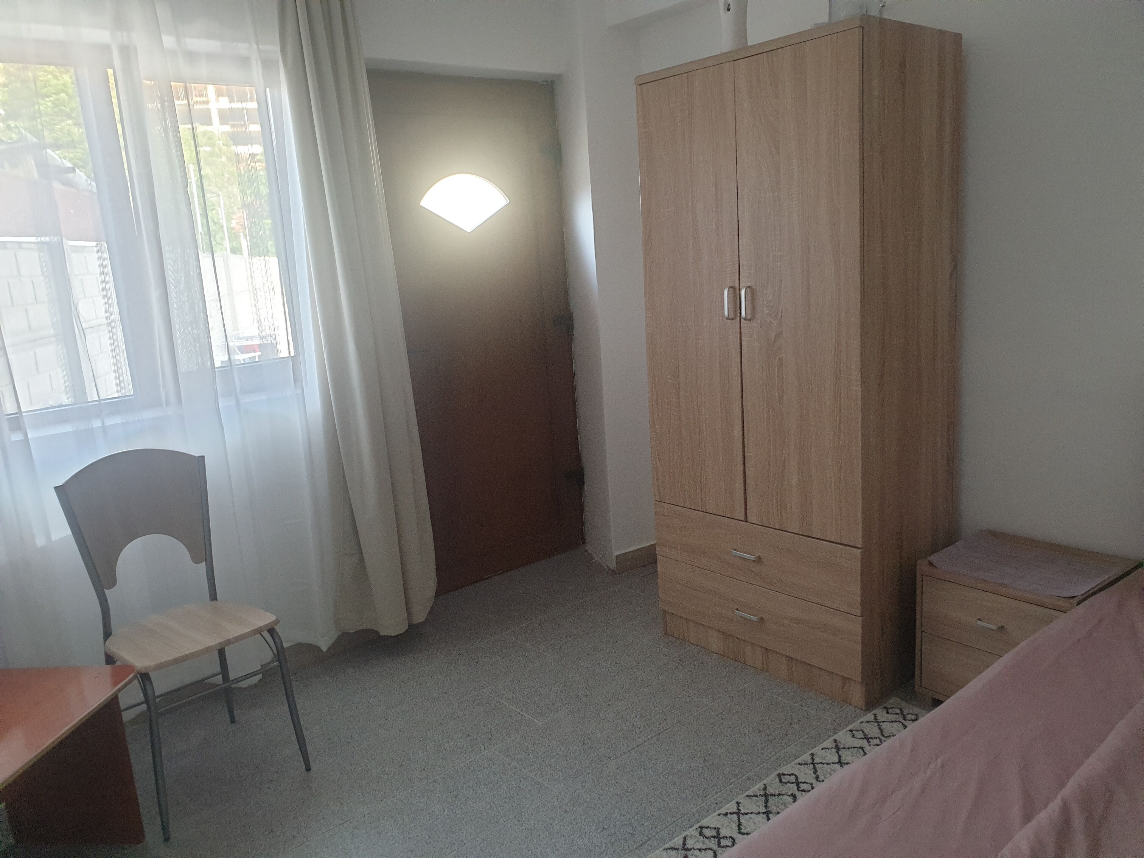 A room with a wooden wardrobe, a chair, a small table, and a window with curtains.