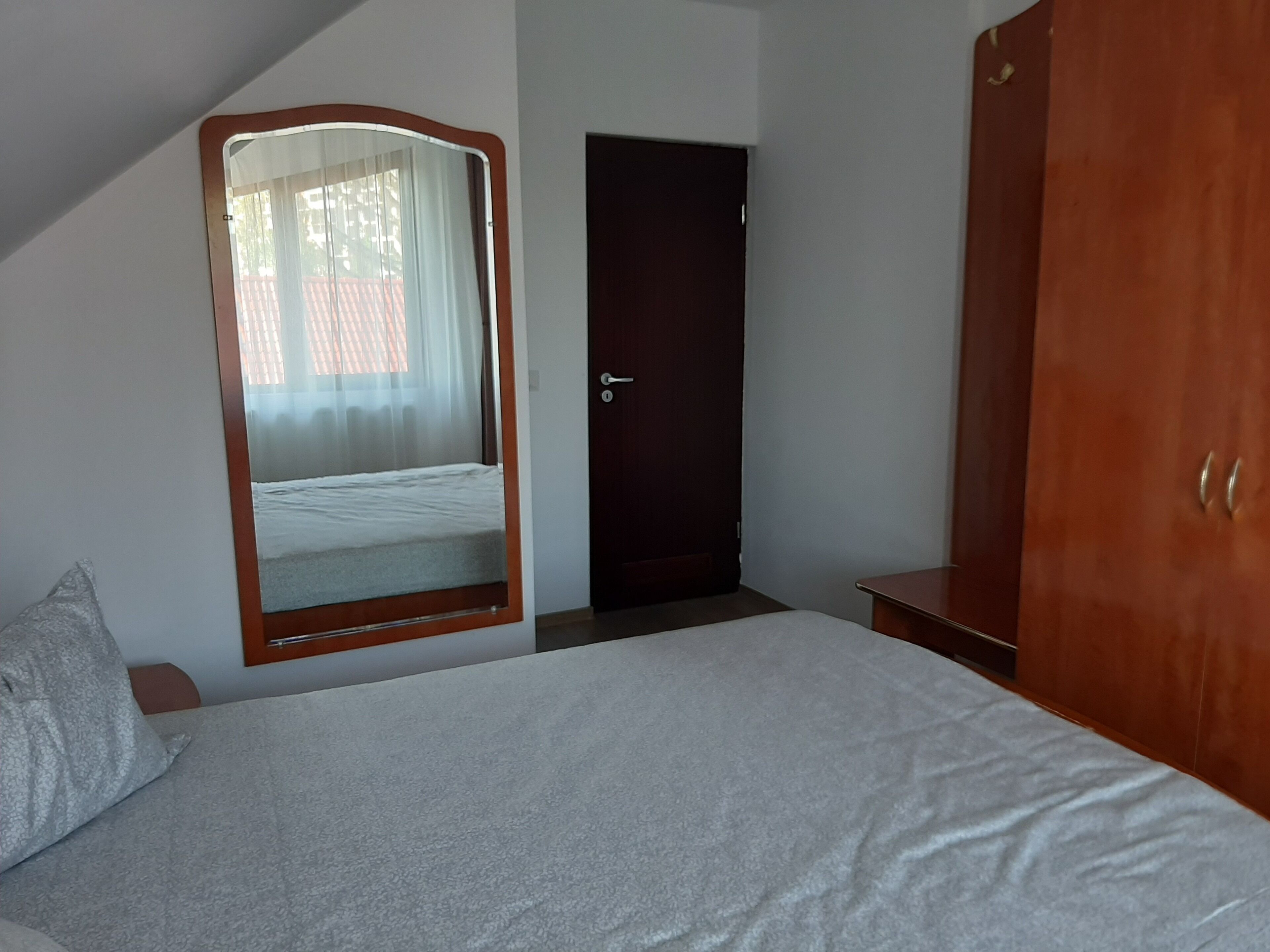 A bedroom with a bed, a wardrobe, a door, and a mirror reflecting a window with curtains.