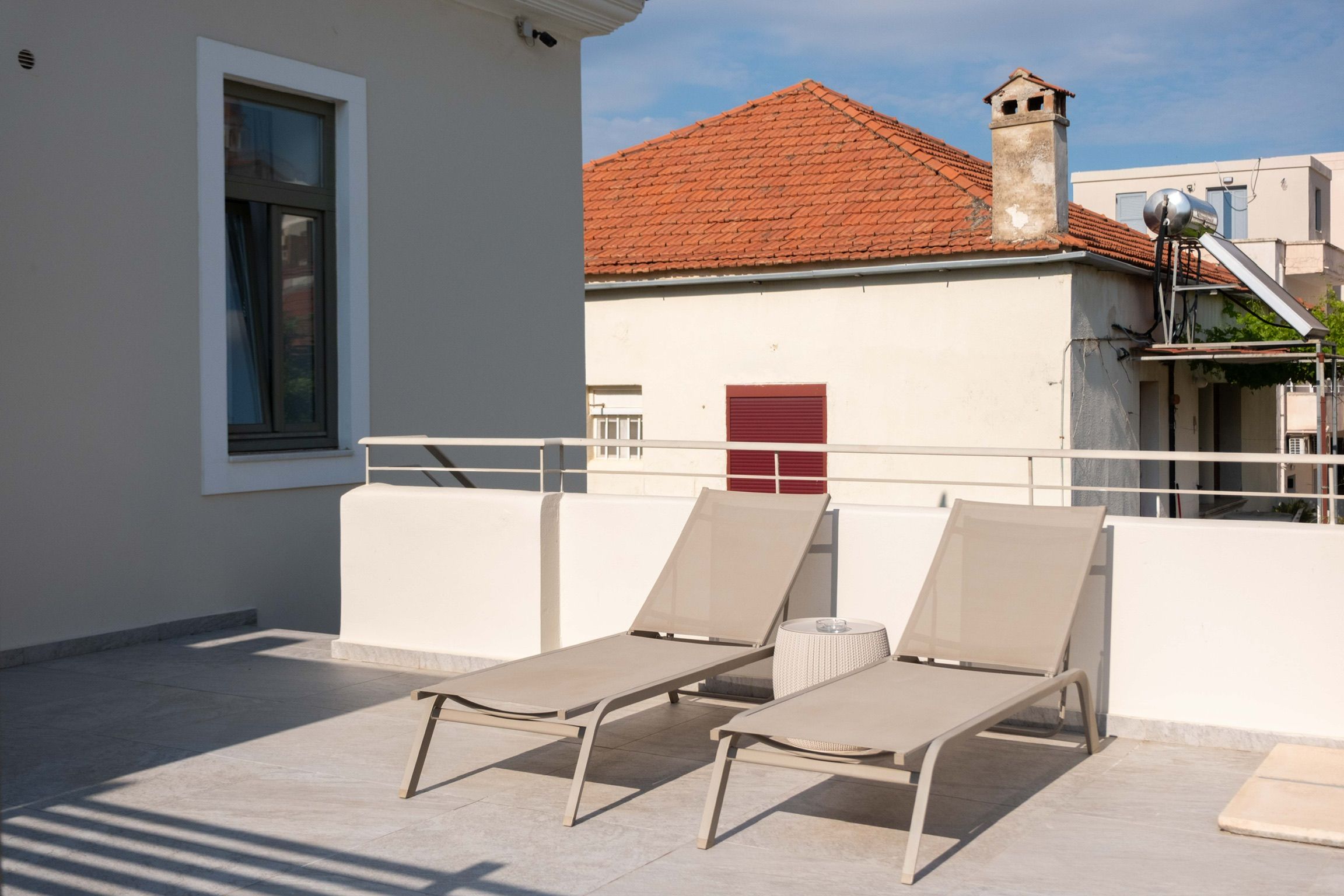 A rooftop terrace with two lounge chairs and a red shuttered window.