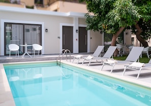 A swimming pool with lounge chairs and a table, surrounded by a building with doors and a tree. - Epavli Boutique Luxury Suites (Chania)