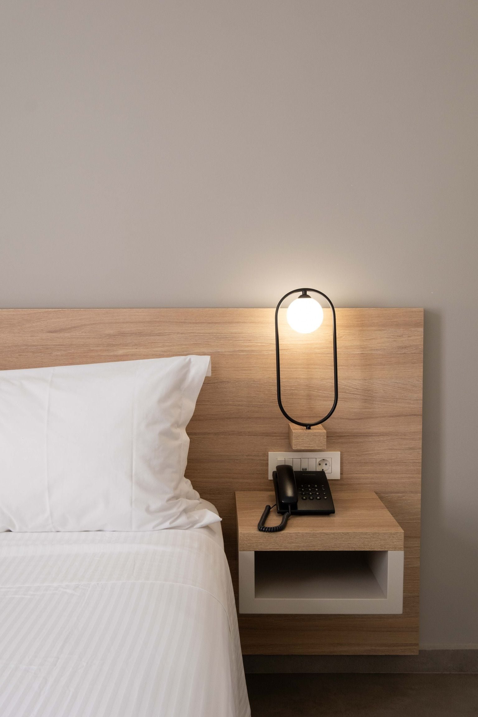 Ein ordentlich bezogenes Bett mit weißen Leinen, ein Nachttisch mit einem schwarzen Telefon und einer wandmontierten Lampe.