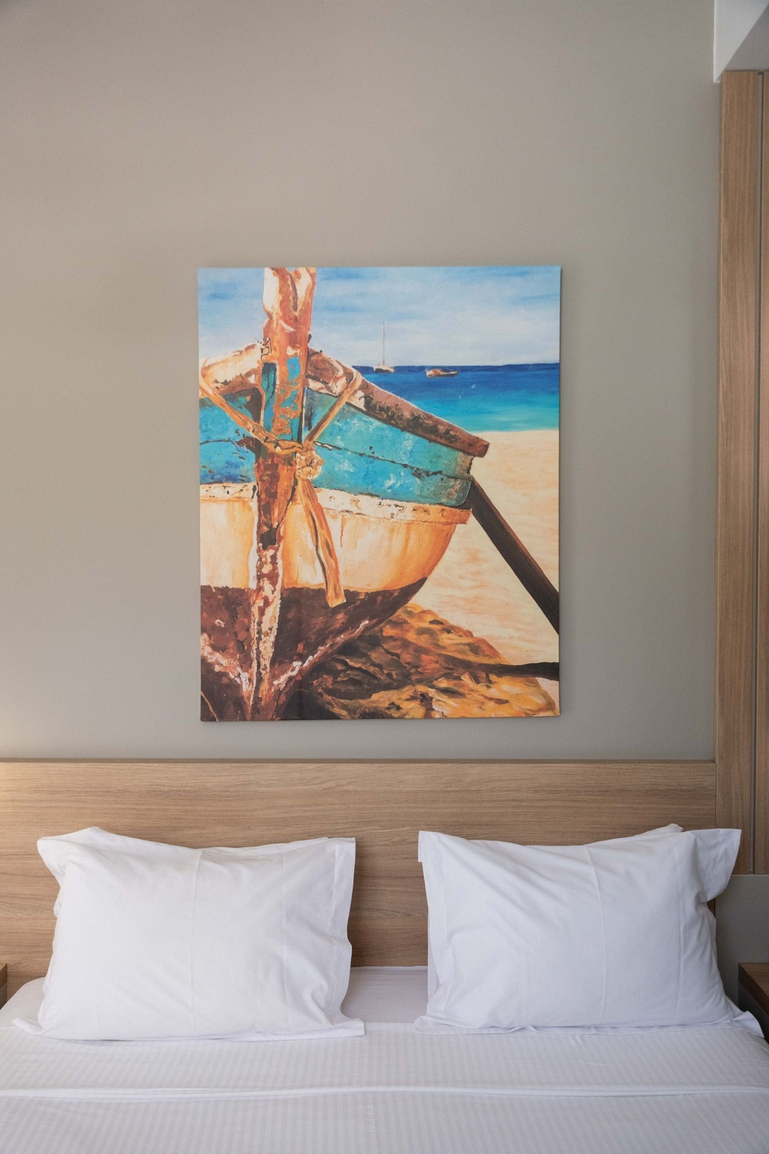 A painting of a boat on a beach, hung on a wall above a bed with white pillows.