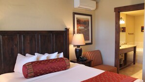 Ein Hotelzimmer mit einem großen Bett, einem Nachttisch, einer Lampe, einem Sessel, einem Bild und einem durchgegangenen Tür erkennbaren Badezimmer.