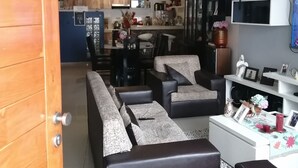 Ein modernes Wohnzimmer mit einer Couch, Sesseln und einem Couchtisch. Im Hintergrund ist eine Küchenzeile mit einem Kühlschrank, einer Mikrowelle und Schränken zu sehen.