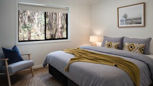 A bedroom with a bed, a chair, a window with a forest view, a bedside lamp, and a framed picture on the wall.