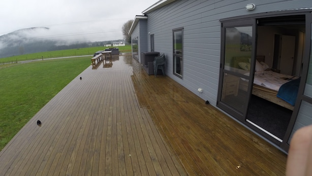 A wooden deck with a table and chairs, a sliding glass door leading to a bedroom, and a view of a green field and distant buildings. - Full holiday home to yourself, up to 21 guests (Atiamuri)