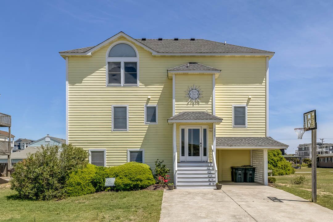 A two-story yellow house with a front porch, a small garage, and a basketball hoop in the driveway.