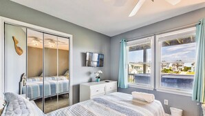 A bedroom with a bed, a dresser, a TV, a window with curtains, and a view of buildings outside. - Sea the Day | 717 ft from the beach | Private Pool, Hot Tub | Nags Head (Nags Head)