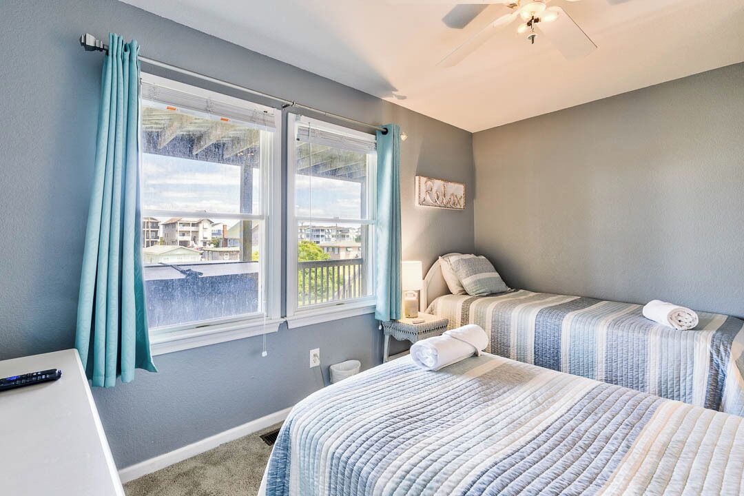 A bedroom with two beds, a window with curtains, a ceiling fan, and a view of buildings outside.