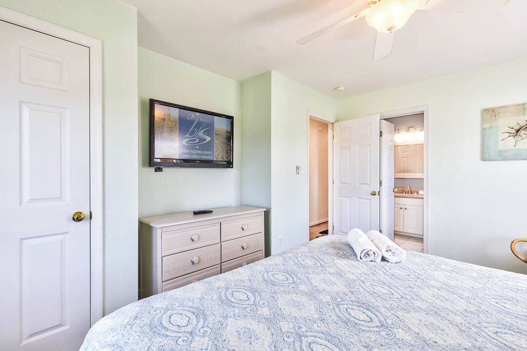 Une chambre à coucher comprenant un lit, une commode, un téléviseur fixé au mur, et une salle de bain visible à travers une porte ouverte.