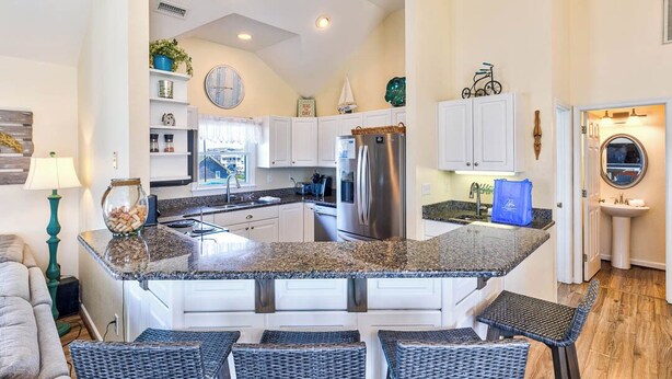 A modern kitchen with granite countertops, white cabinetry, and a stainless steel refrigerator. There are bar stools and a small island with a sink. - Sea the Day | 717 ft from the beach | Private Pool, Hot Tub | Nags Head (Nags Head)