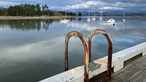 Un lago tranquilo con varias embarcaciones ancladas, un muelle de madera con barandillas de metal oxidadas y una orilla boscosa bajo un cielo nublado.