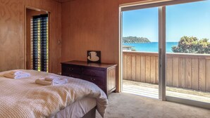 A bedroom with a large bed, a wooden dresser, and a sliding glass door leading to a balcony with a view of the sea. - SANDCASTLE ON ONETANGI | Be My Guest (Onetangi)