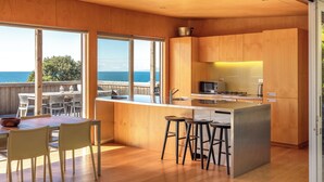 A modern kitchen with wooden cabinets, a large island with stools, and a view of the ocean. - SANDCASTLE ON ONETANGI | Be My Guest (Onetangi)