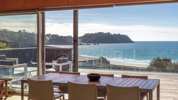 A wooden deck with a table and chairs, overlooking a beach with clear blue water and a forested island. - SANDCASTLE ON ONETANGI | Be My Guest (Onetangi)