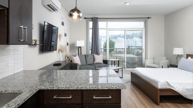 A modern hotel room with a kitchen area, a large bed, a sofa, and a view of the city. - Studio King Suite w/ Kitchen | 201 (Asheville)