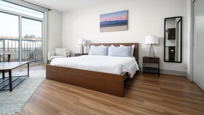 A modern bedroom with a large bed, a desk, and a chair. There is a large window and a painting on the wall. - Studio King Suite w/ Kitchen | 201 (Asheville)