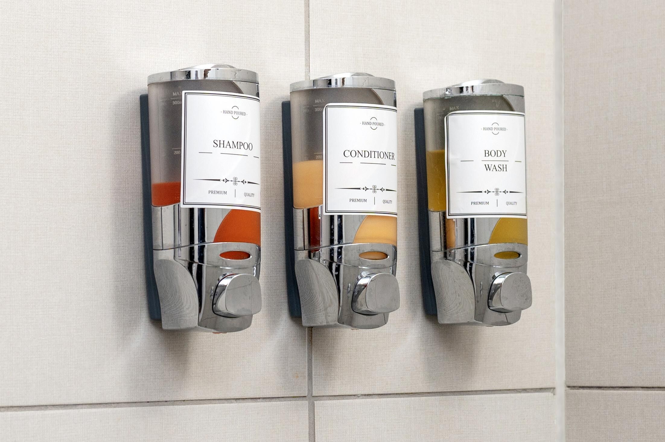 Three hand-poured dispensers for shampoo, conditioner, and body wash in a bathroom.