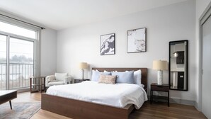 Ett modernt hotellrum med en stor säng, två sängbord med lampor, en stol och ett stort fönster med utsikt över staden.