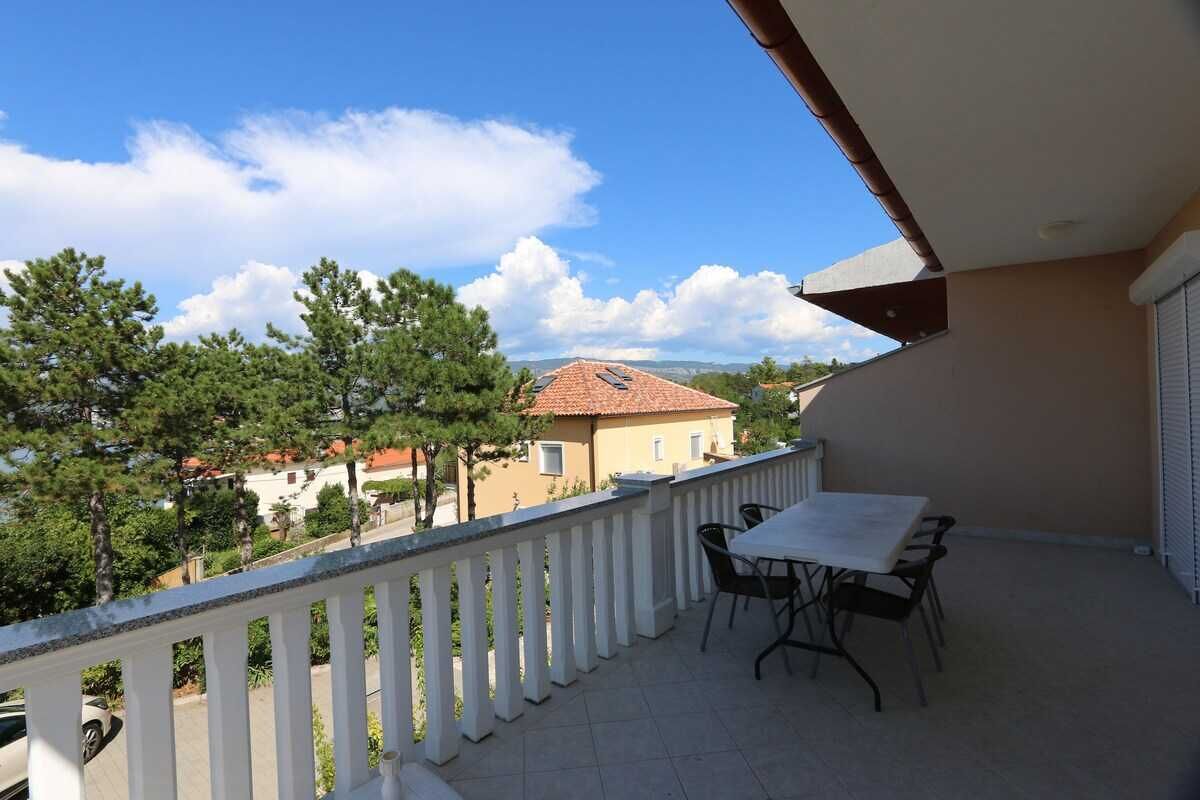 A balcony with a table and chairs, a white railing, and a view of houses and trees.
