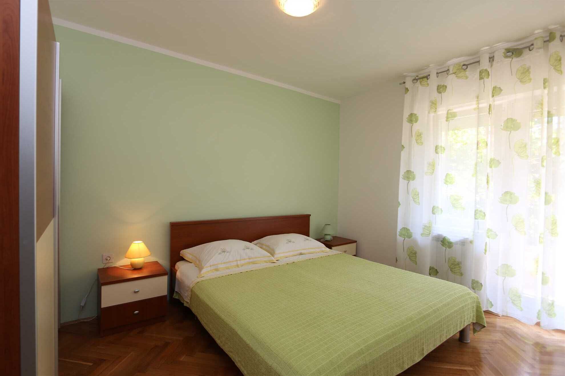 Ein Schlafzimmer mit Bett, Nachttisch, Lampe und einem Fenster mit leichtem Vorhang.