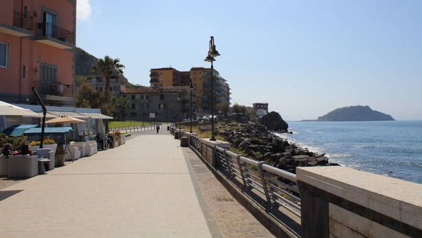 A seaside promenade with outdoor seating, a stone railing, and buildings in the background. - B&B Allerìa (Pozzuoli)
