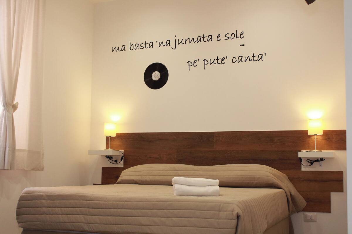 Una cama bien hecha con cabecera, dos lámparas de noche y una pared con un disco y texto.