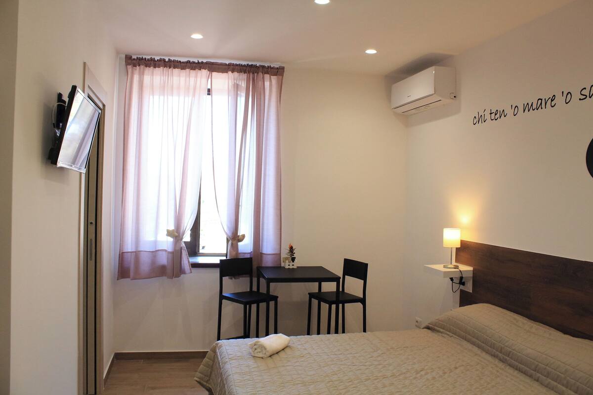 A hotel room with a bed, a desk with two chairs, a TV, and an air conditioner.