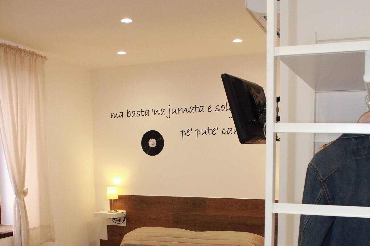A bedroom with a bed, a nightstand, a wall-mounted TV, and a shelf with a jacket.