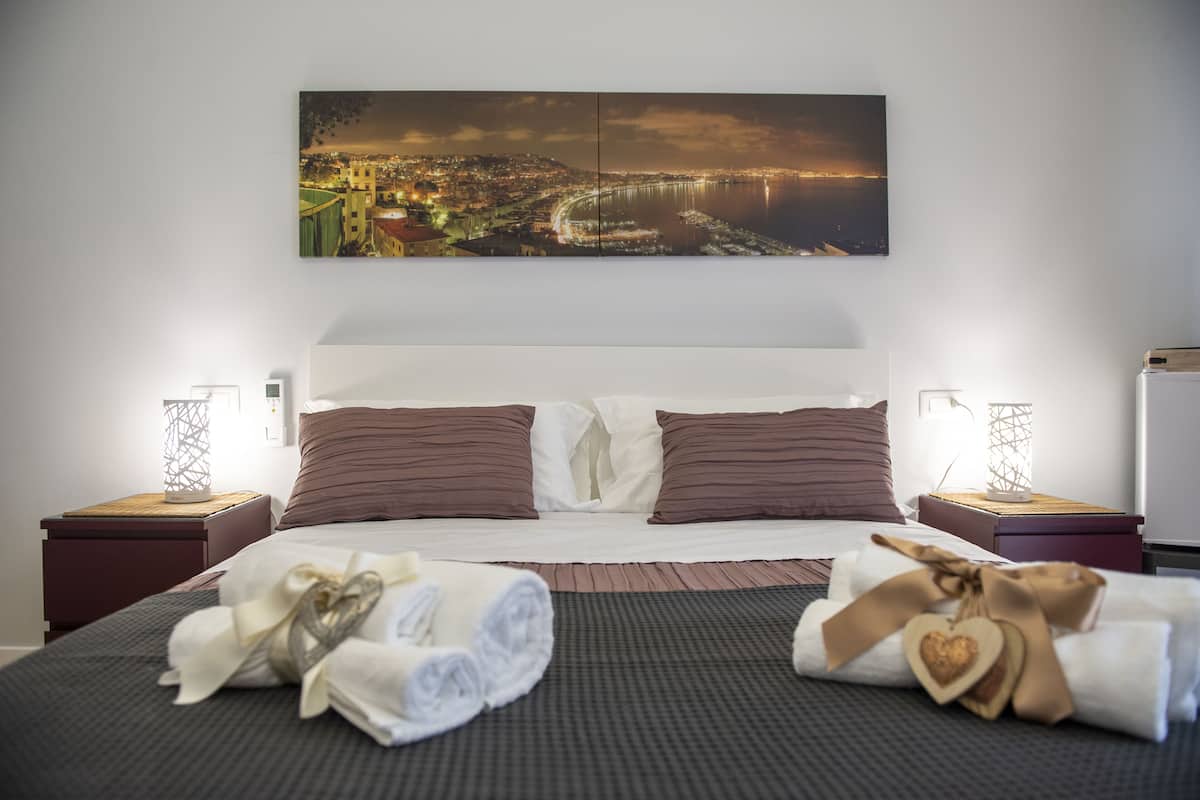 Una cama bien hecha con ropa de cama marrón y blanca, dos mesitas de noche con lámparas y un cuadro de paisaje urbano sobre la cama.