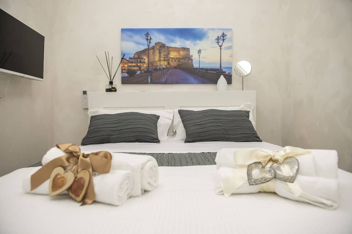 Habitación de hotel con cama, un cuadro de un castillo, un televisor y una mesita de noche con lámpara y una botella.