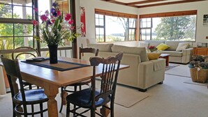 A dining area with a wooden table and chairs, a vase of flowers, and a view of the outdoors through large windows.