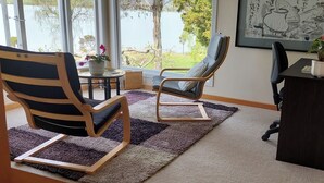 A room with a large window, a wooden chair, a cushioned chair, a small table with a potted plant, and a framed artwork on the wall.