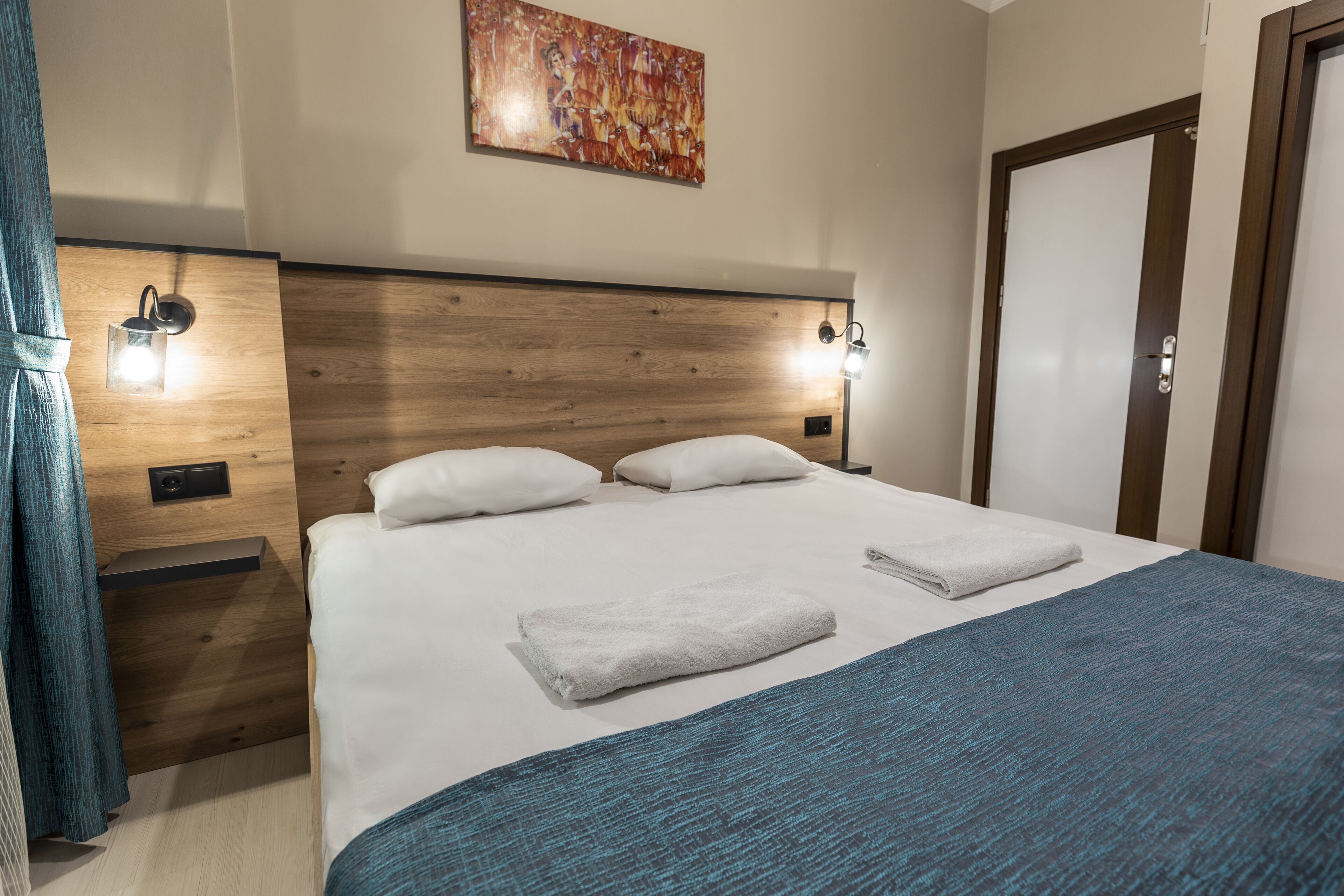 Una cama bien hecha con sábanas blancas y una manta azul, un cabecero de madera, dos luces de lectura fijadas a la pared y una puerta a la derecha.