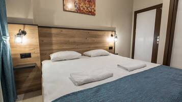 Una cama bien hecha con sábanas blancas y una manta azul, un cabecero de madera, dos luces de lectura fijadas a la pared y una puerta a la derecha.