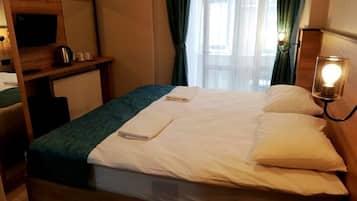 A hotel room with a wooden bed, a bedside table with a lamp, a TV, and a view of the outdoors through sheer curtains.