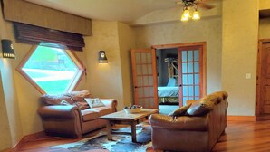 Een gezellige woonkamer met een bruine leren bank, een houten salontafel en een groot raam met uitzicht op de natuur.