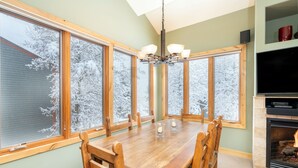 A dining room with a wooden table and chairs, large windows with snow-covered trees, a fireplace, and a television on a shelf. - Beautiful 3 bedroom Condo close to Lift 7 (Telluride)