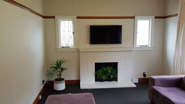 A living room with a fireplace, a flat-screen TV mounted above it, two windows with white frames, and a potted plant. - right in the town centre (Whakatane)