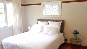 A neatly made bed with white linens in a room with a wooden floor, a window with white curtains, and a small round table with a vase of flowers. - right in the town centre (Whakatane)