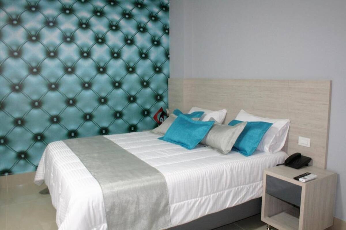 A neatly made bed with white bedding and blue pillows, a wooden headboard, and a bedside table with a phone and remote control.