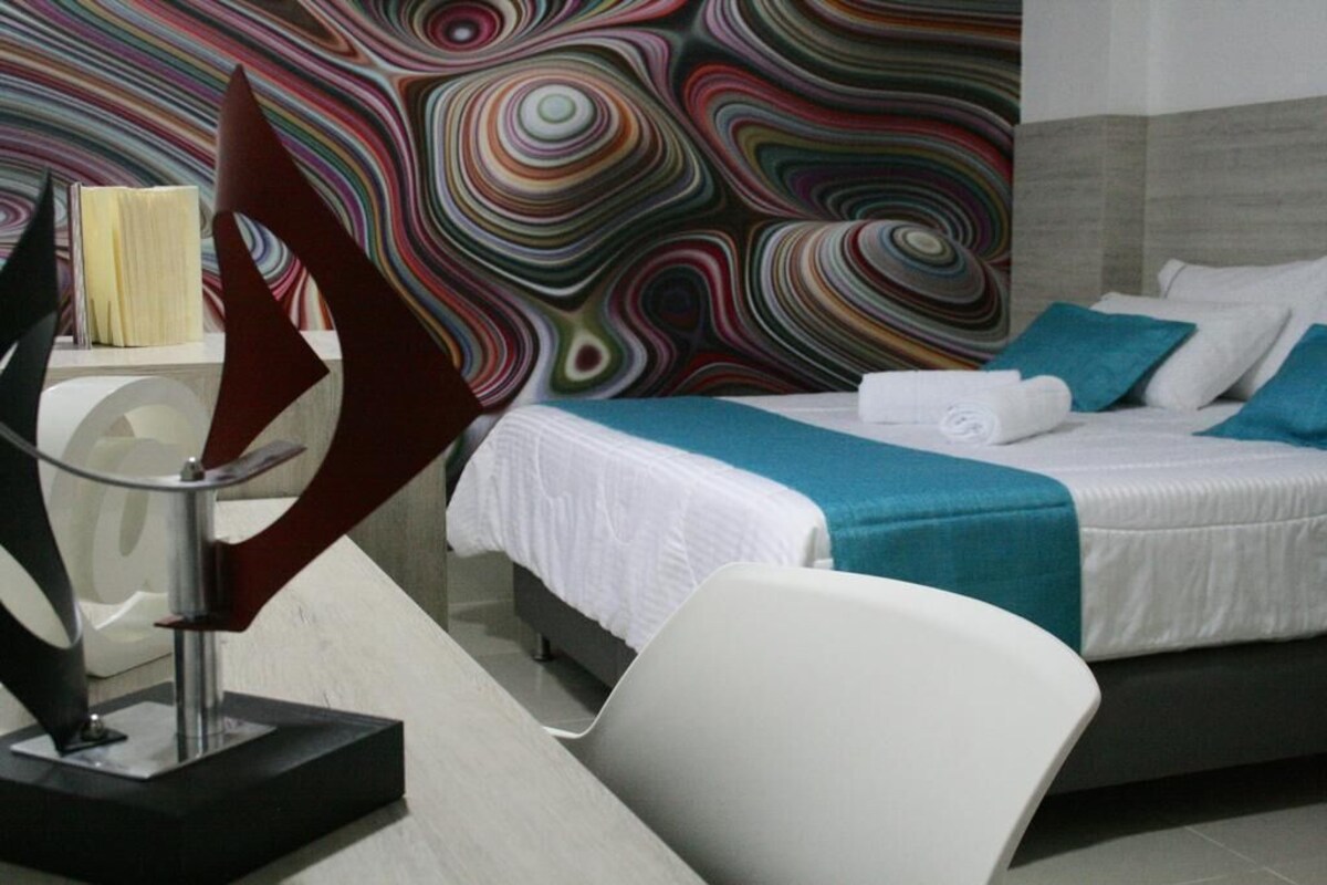 Une chambre d’hôtel moderne avec un lit, une chaise blanche et un motif mural abstrait aux couleurs vives.