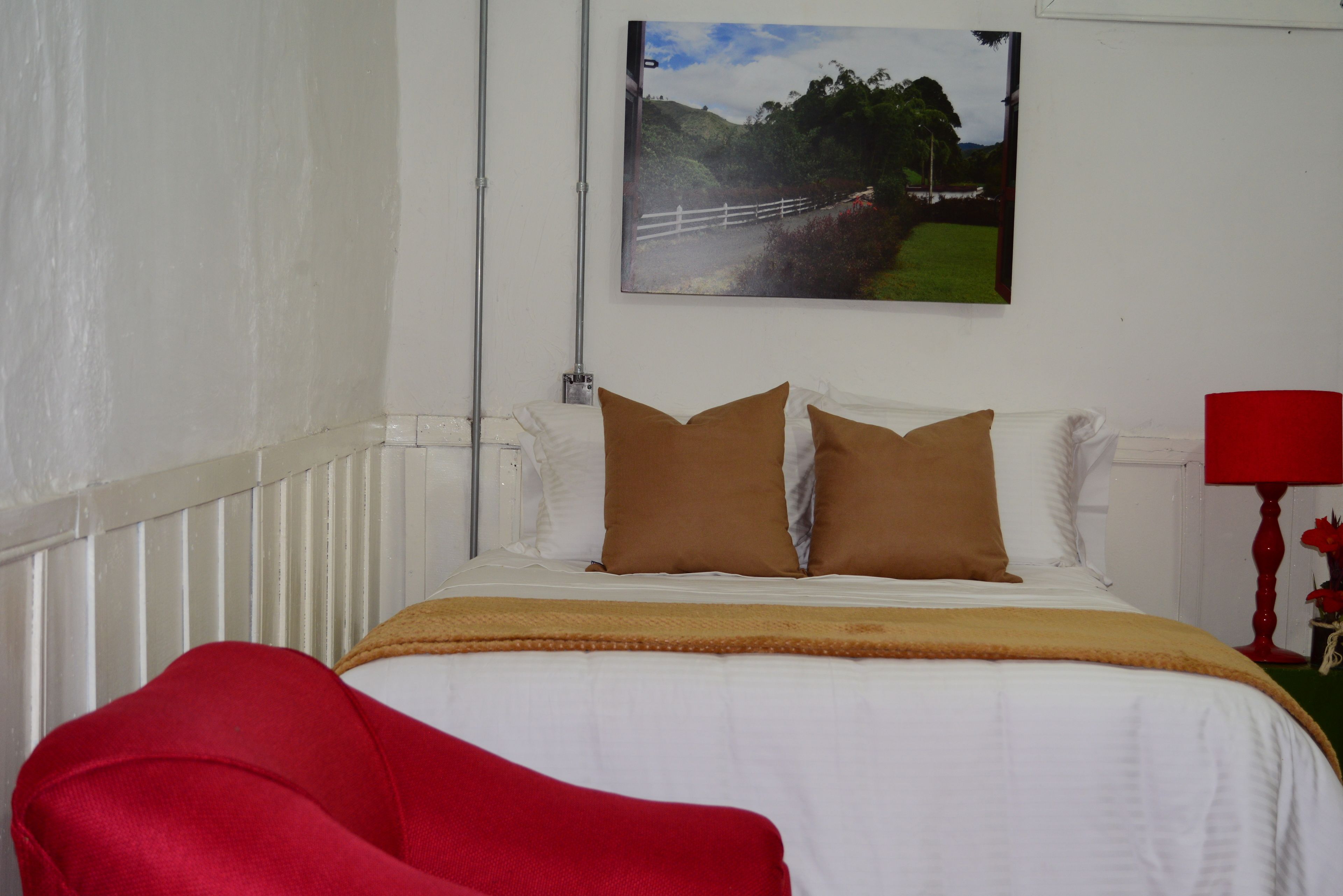 A bedroom with a white bed, two brown pillows, a red chair, and a painting of a landscape on the wall.