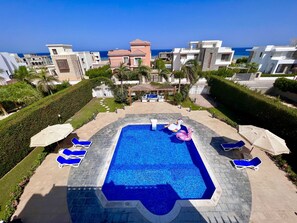 A swimming pool with lounge chairs and a decorative swan float, surrounded by well-maintained hedges and a view of the sea.