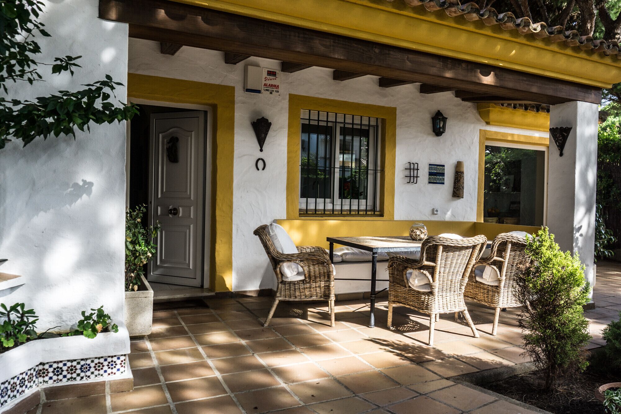 Une terrasse extérieure avec une table et des chaises en osier, une porte et une fenêtre avec des vitres à croisillons.
